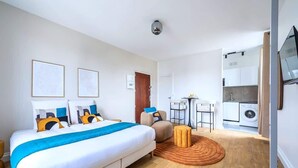 A modern bedroom with a large bed, a small seating area, and a kitchenette in the background. - Studio chaleureux au coeur de ville Issy (Issy-les-Moulineaux)