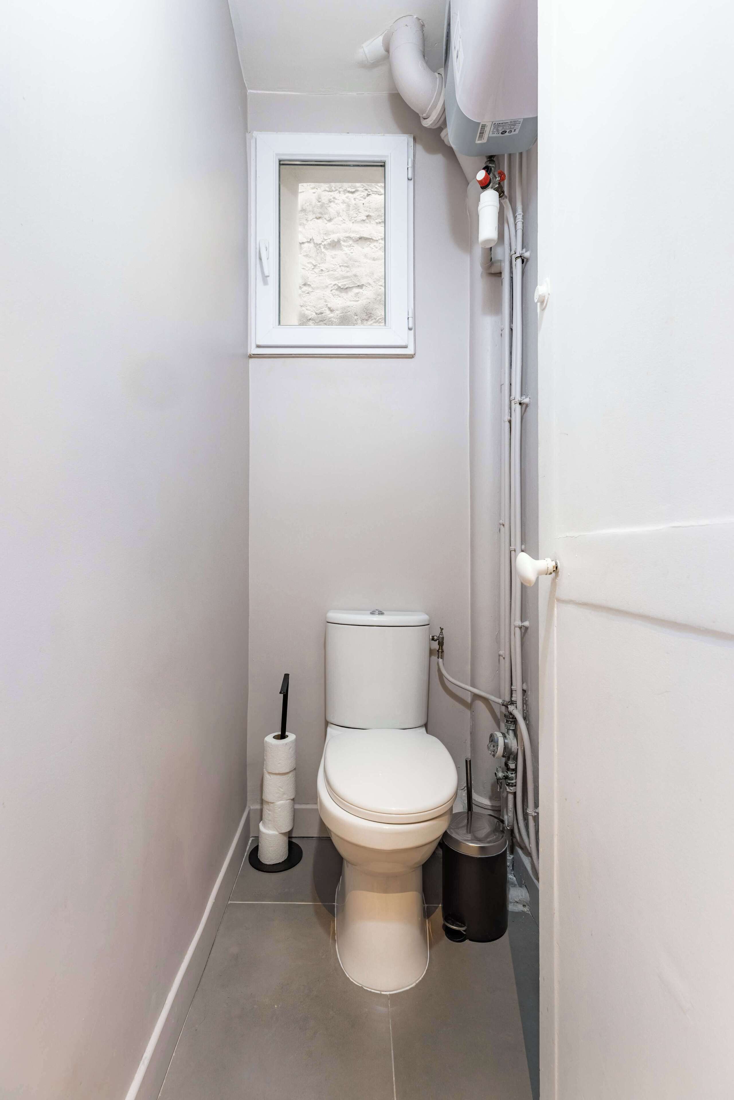 A compact bathroom with a toilet, a small window, and a trash bin.