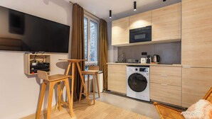 Une cuisine compacte équipée d’un four à micro-ondes, d’un four et d’un lave-linge. On y trouve un coin repas en bois avec deux chaises et un téléviseur fixé au mur.