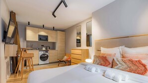 Une chambre d’hôtel moderne dotée d’un grand lit, d’un coin salon avec un fauteuil, d’une petite cuisine équipée d’un lave-linge et d’un miroir au mur.