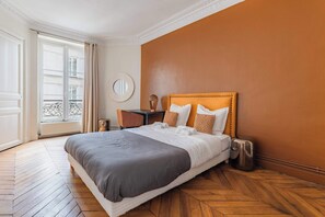 A hotel room with a large bed, a wooden floor, a round mirror, and a view of a building through the window. - Grand appartement 80m2 2BR - Beaubourg / Notre-Dame (Paris)