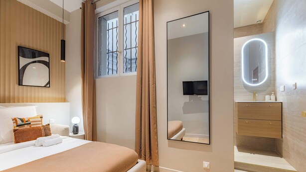 A modern hotel room with a large bed, a bedside table with a lamp, a mirror with a lighted frame, a TV, and a bathroom with a vanity and a toilet. - Élégant 3 pièces climatisé au cœur de Paris 10 (Paris)