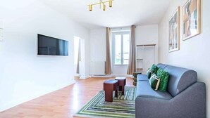 Un salon moderne avec un canapé gris, deux petites tables d’appoint rondes, un téléviseur fixé au mur et un tapis à motifs sur le plancher en bois.