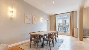 Een ruime kamer met een grote eettafel en stoelen, een bank en uitzicht op een balkon met hekwerk.