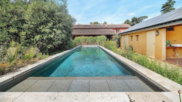 Ein rechteckiger Pool mit Steinrand, umgeben von Grünpflanzen und einem Gebäude mit Ziegeldach im Hintergrund.