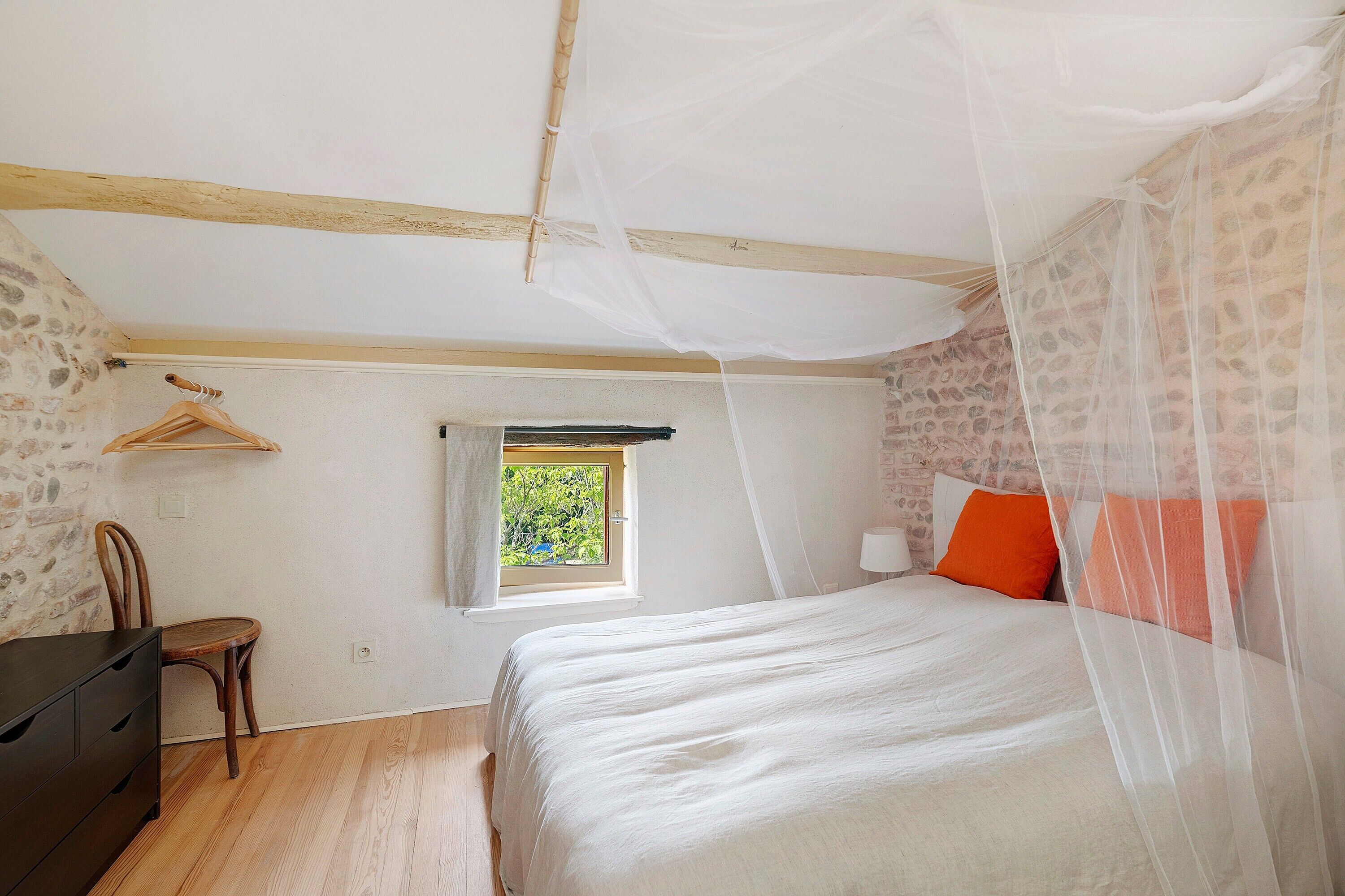 A bedroom with a bed, a chair, a desk, a window, and a mosquito net.