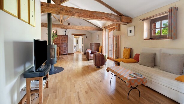 A spacious living room with wooden beams, a sofa, a television on a stand, and a table with a tablecloth.