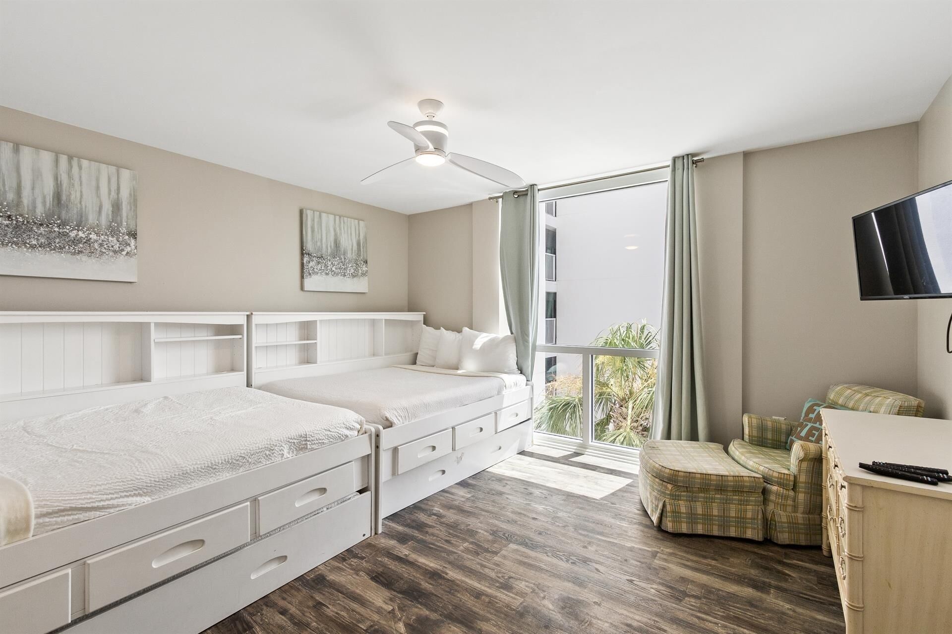 Une chambre avec deux lits, un ventilateur de plafond, une télévision et un balcon avec une chaise.