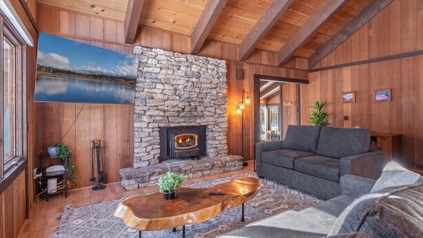 A cozy living room with a stone fireplace, wooden beams, a gray sofa, and a large window overlooking a lake. - Dollar Point - Observation Dr. 3 Bedroom Home (Tahoe City)