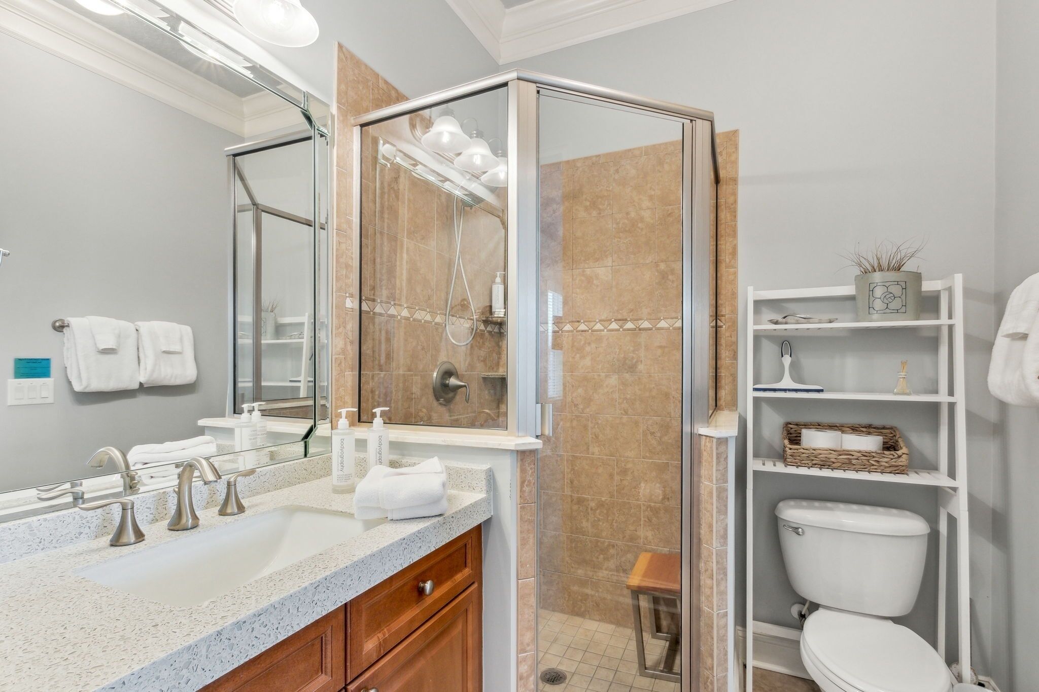 A modern bathroom with a glass shower enclosure, a large mirror, a sink with a granite countertop, and a toilet with a basket shelf.