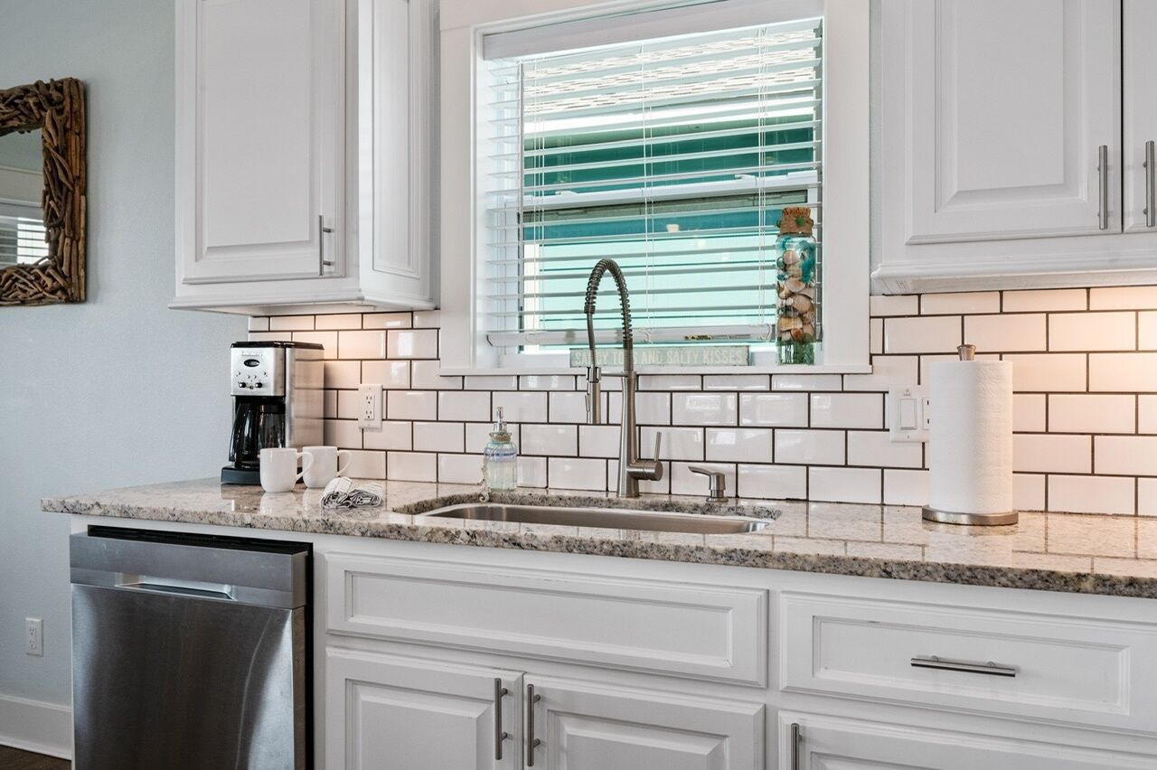 A kitchen with white cabinets, a stainless steel dishwasher, a sink with a modern faucet, and a window with blinds.