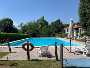 A rectangular swimming pool with a wooden deck, surrounded by white lounge chairs and umbrellas, with a hedge and trees in the background. - Casa Valentina - Pool, Tennis & AC - Lake Maggiore (Montegrino Valtrvaglia)
