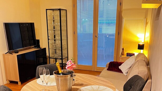 Una sala de estar moderna con una mesa redonda de madera, dos sillas grises, un sofá, un televisor y un aparador de vidrio con copas de vino.