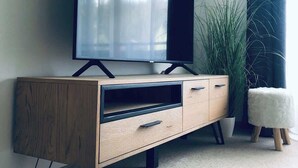 A modern living room with a flat-screen TV on a wooden TV stand, a potted plant, and a fluffy white ottoman. - Wanderlust Chiemgau (Bad Endorf)