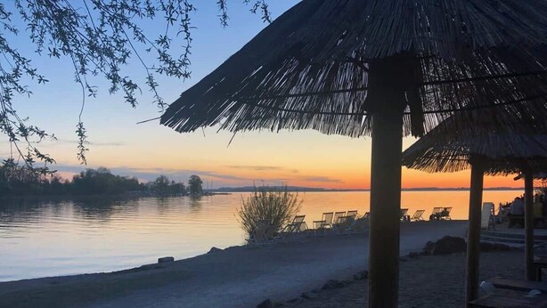 A beach with a thatched-roof structure, a calm body of water, and a sunset in the background. - Wanderlust Chiemgau (Bad Endorf)