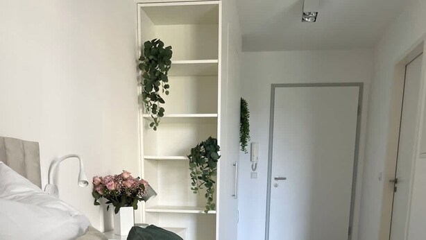 Une chambre à coucher moderne avec une couvre-lit matelassé blanc, une tête de lit blanche et une table de chevet ornée d’un vase de fleurs.