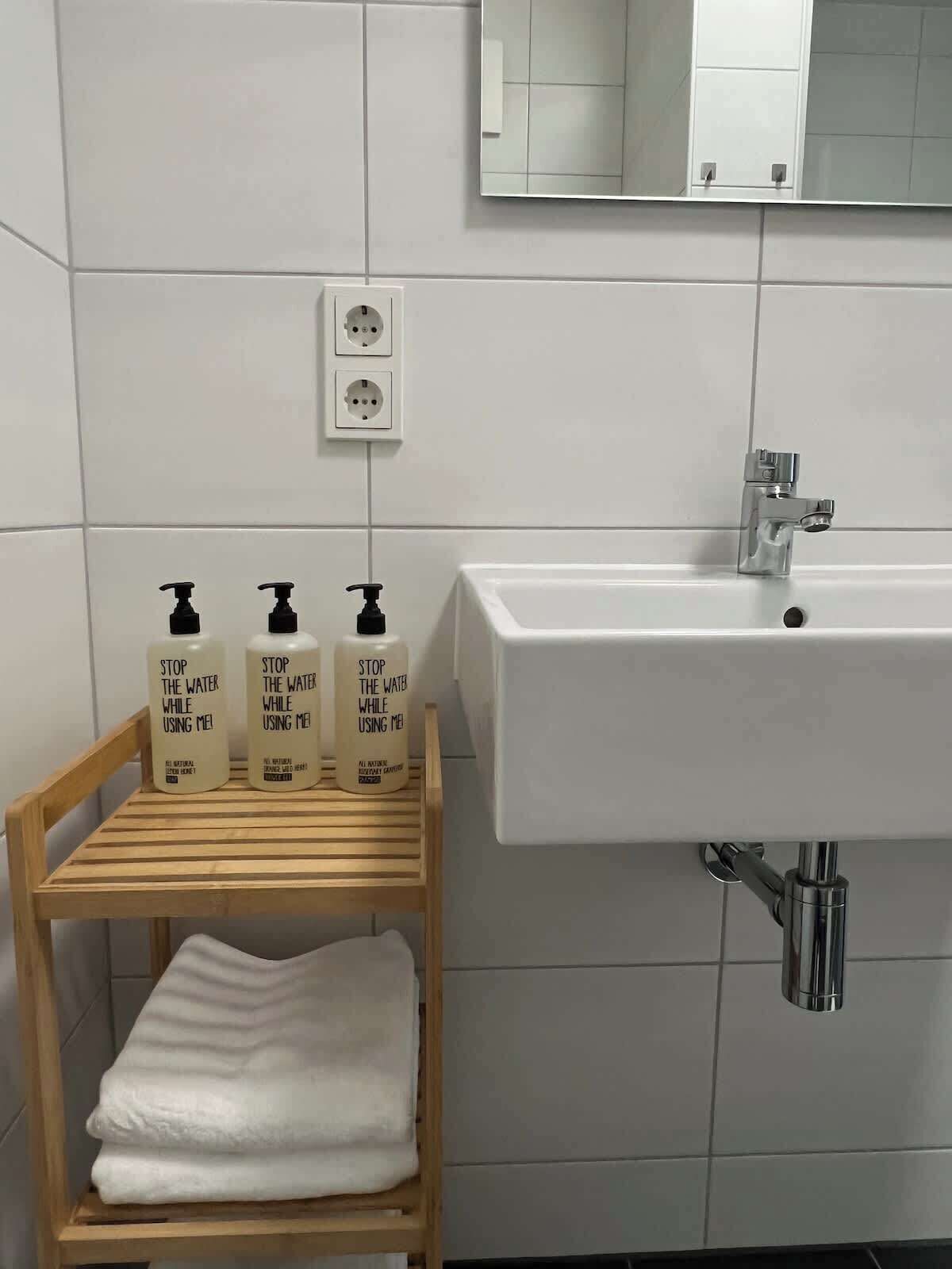 A bathroom with a white sink, a mirror above it, and three bottles of soap on a wooden shelf.