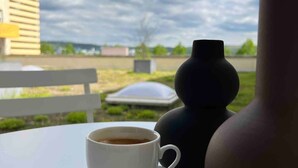 Une tasse de café blanche, posée sur une soucoupe blanche, sur une table. En arrière-plan, on aperçoit un vase foncé et un bâtiment.