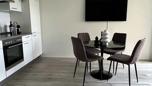 Une cuisine moderne avec des meubles blancs, une cuisinière noire et une machine à café. On y trouve un coin repas avec une table ronde, quatre chaises et un téléviseur à écran plat fixé au mur.