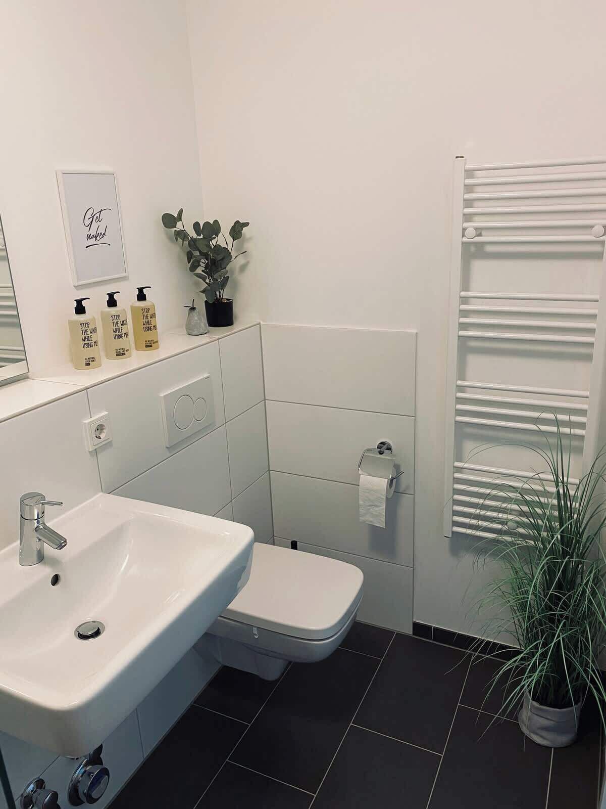 Een moderne badkamer met een witte wasbak, toilet en handdoekhouder. Er staan drie flessen handzeep op de muur, een potplant en een lijst met een bord.