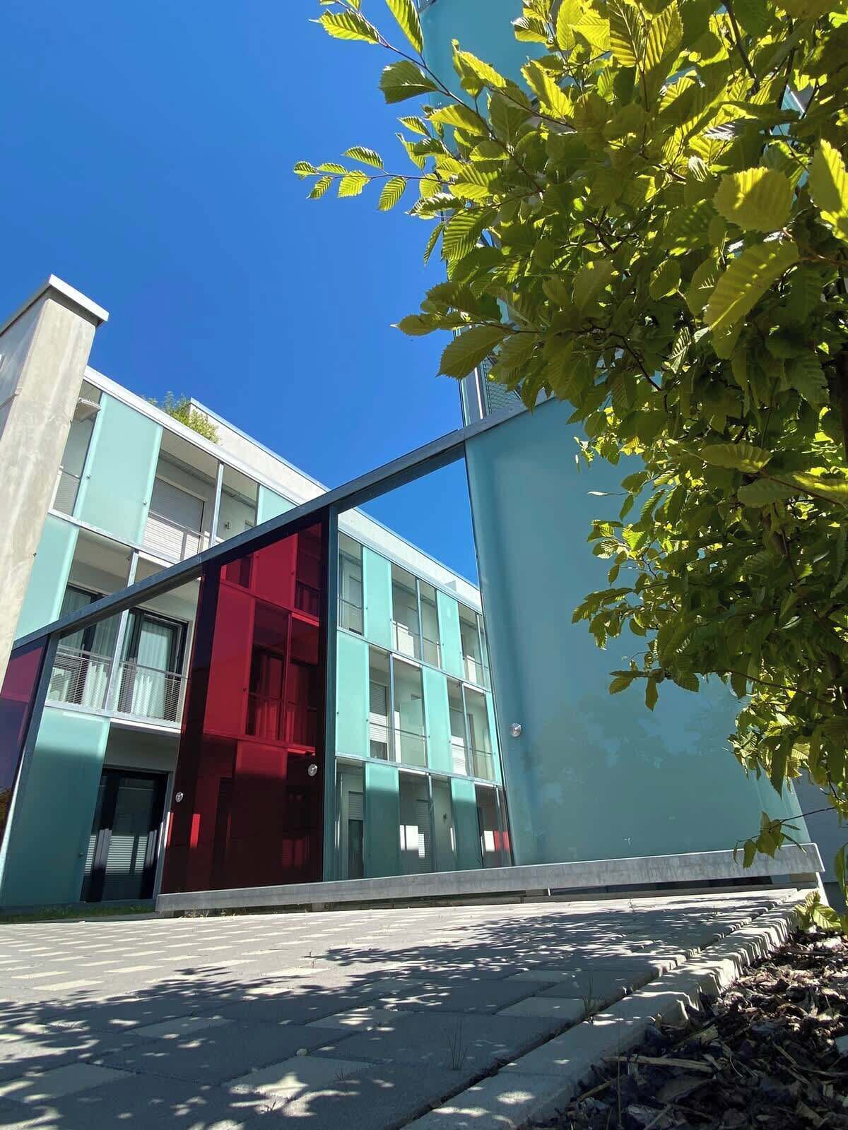A modern building with a glass facade and a red section, featuring a flat roof and a paved ground.