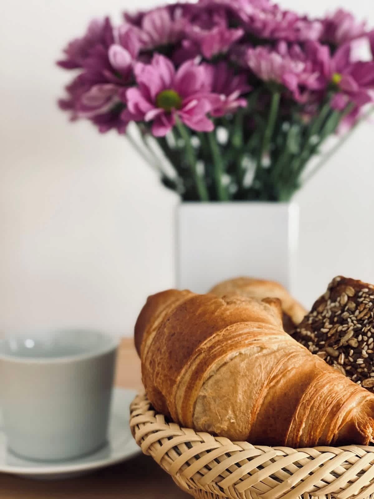 Ein Korb mit einem Croissant, einem Schokoladen-Croissant, einer Tasse und einem Strauß lila Blumen.