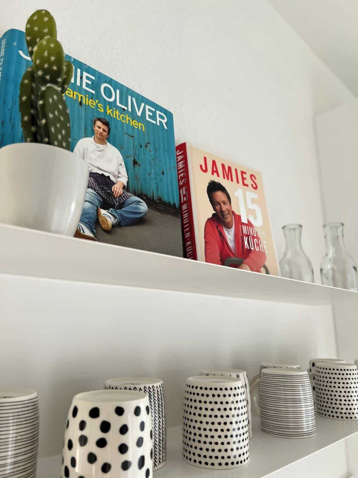 Una estantería con dos libros titulados 'Jamie Oliver's Kitchen' y 'Jamie 15 Minuten Koch'. Debajo de los libros, hay varias tazas blancas con diseños negros.