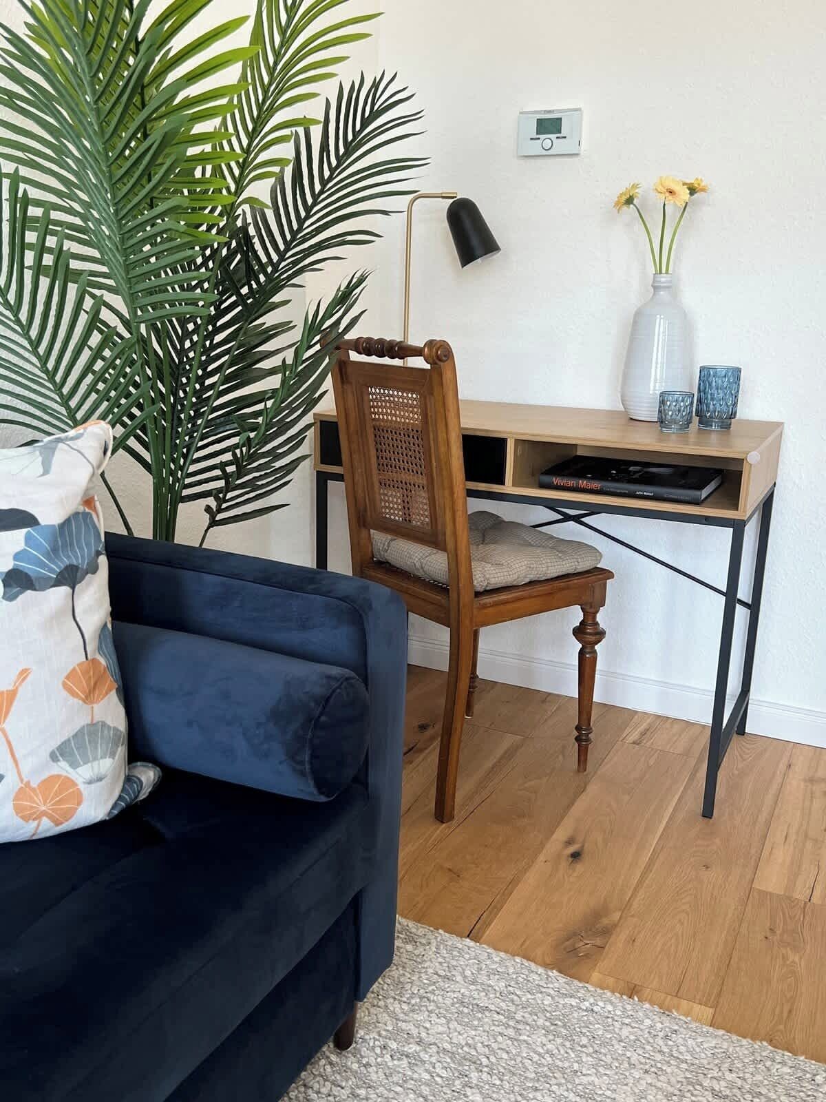 Een kamer met een donkerblauwe bank, een houten stoel, een bureau met een laptop, een potplant en een vaas met bloemen.