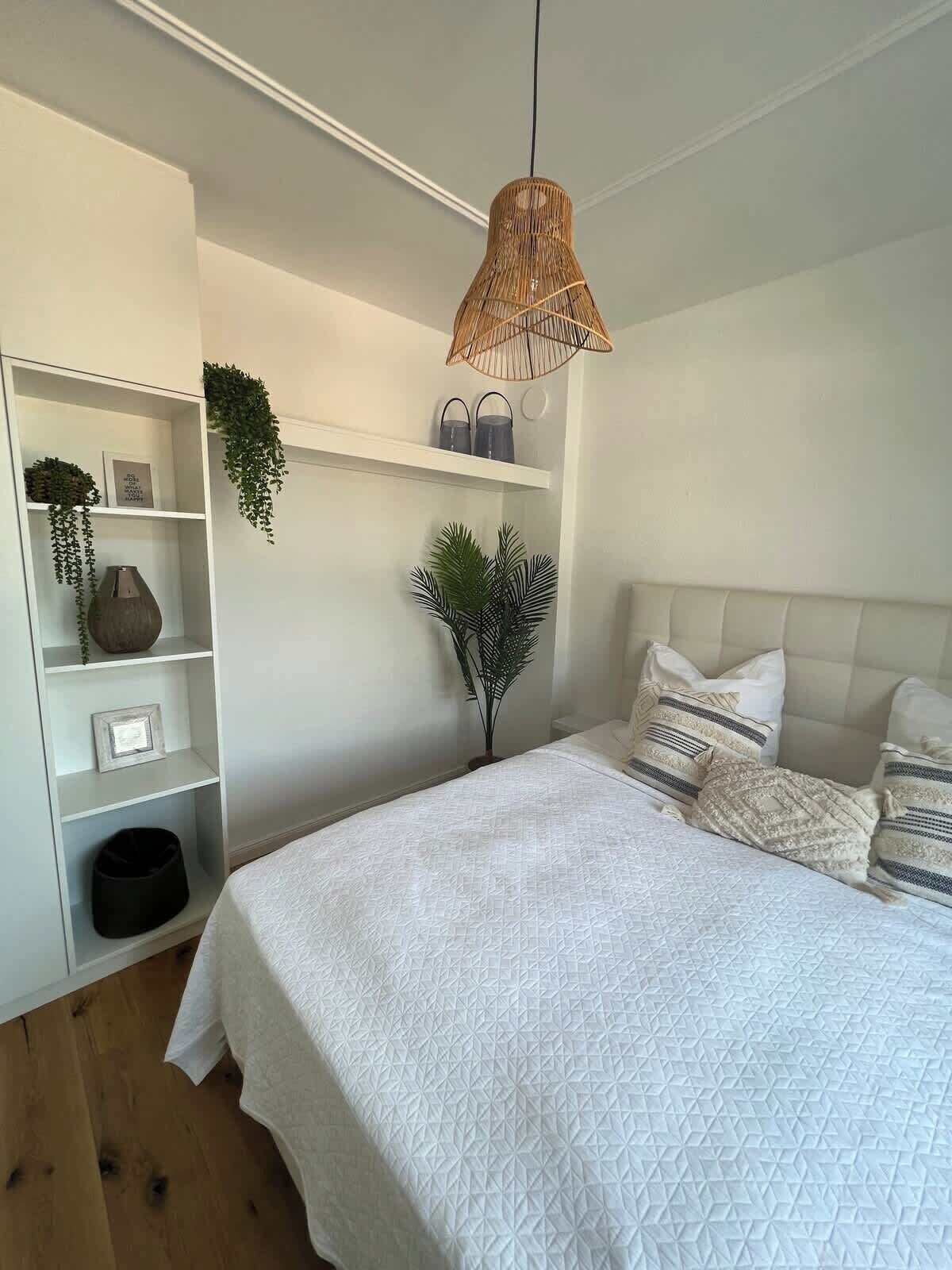Un dormitorio con cama, paredes blancas, una lámpara colgante de ratán y un estante blanco con objetos decorativos.