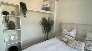 Un dormitorio con cama, paredes blancas, una lámpara colgante de ratán y un estante blanco con objetos decorativos.