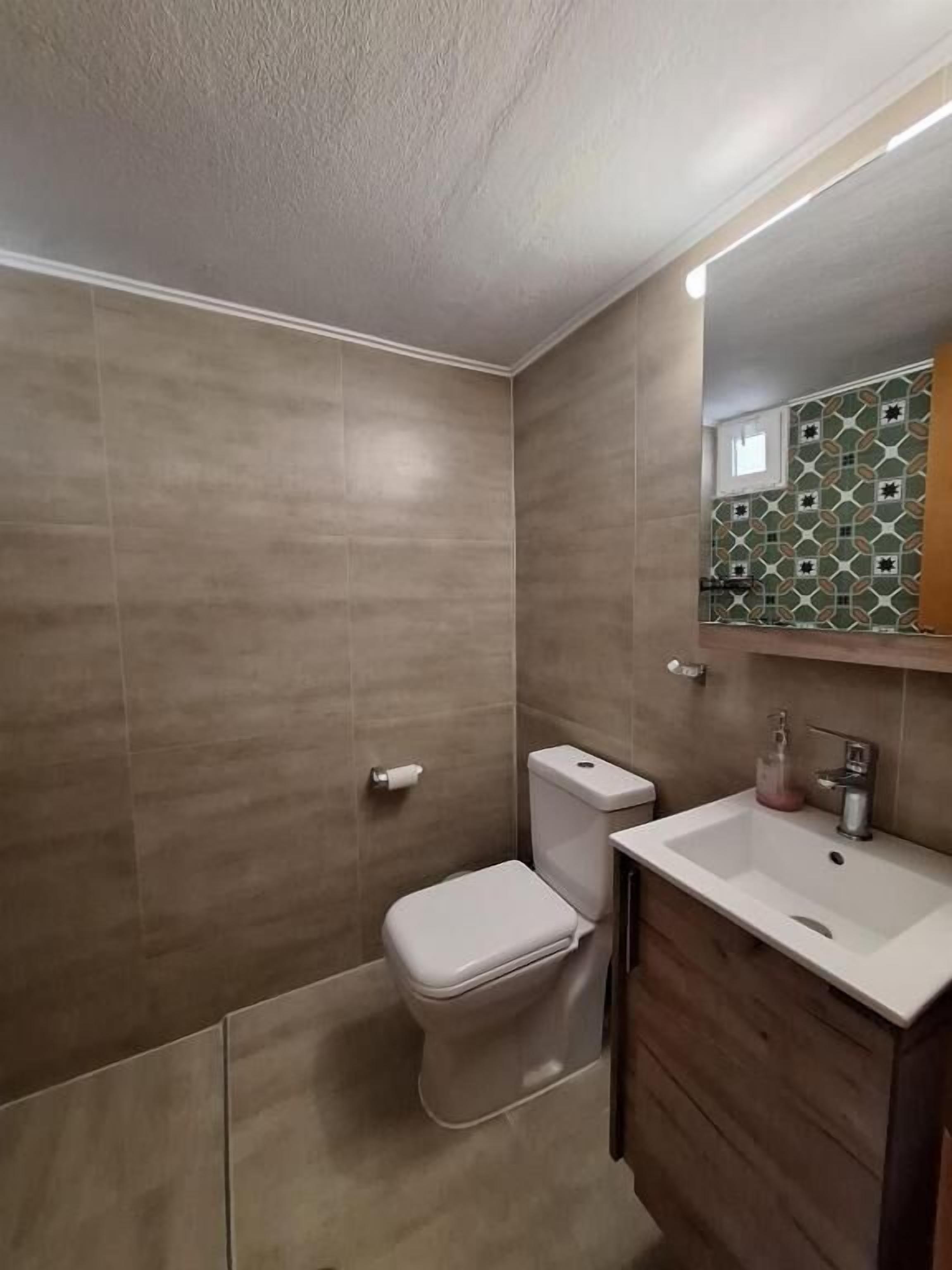 A bathroom with a toilet, sink, and mirror. The walls are tiled, and there is a window above the sink.