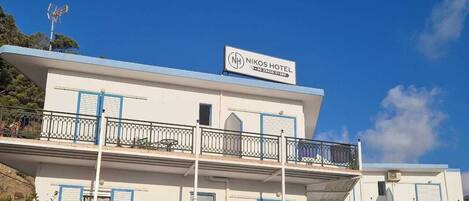 Een meerlaags hotel met balkons, een bord met daarop 'NIKOS HOTEL', en een helderblauwe lucht.