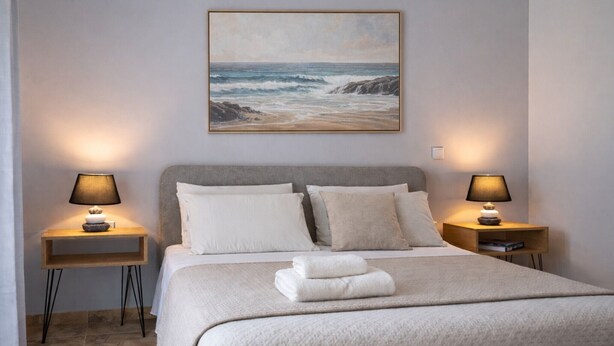 Una camera da letto con un letto, due comodini con lampade, un quadro raffigurante una spiaggia e un letto rifatto con asciugamani piegati.