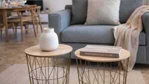 Un soggiorno con un divano grigio, una coperta beige, un vaso bianco e un libro intitolato 'Mediterranean Living' su un tavolino rotondo in legno.