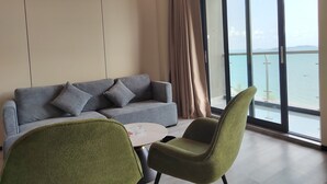 Ett modernt vardagsrum med en grå soffa, gröna fåtöljer, ett litet runt bord och ett stort fönster med utsikt över havet.