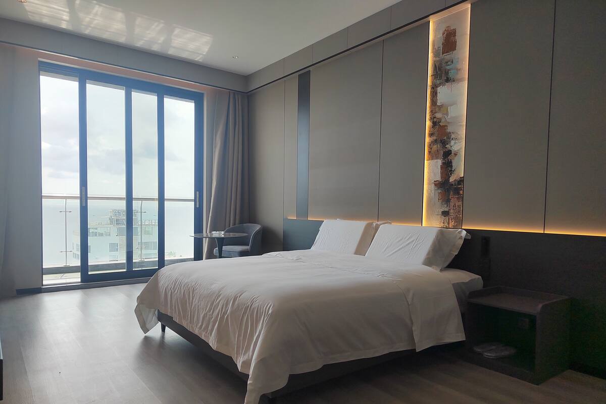 A modern hotel room with a large bed, a small table, and a view of the city through floor-to-ceiling windows.