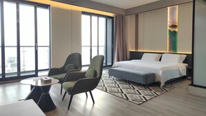 Ett modernt hotellrum med en stor säng, två gröna fåtöljer, ett litet runt bord och utsikt över staden genom stora fönster.