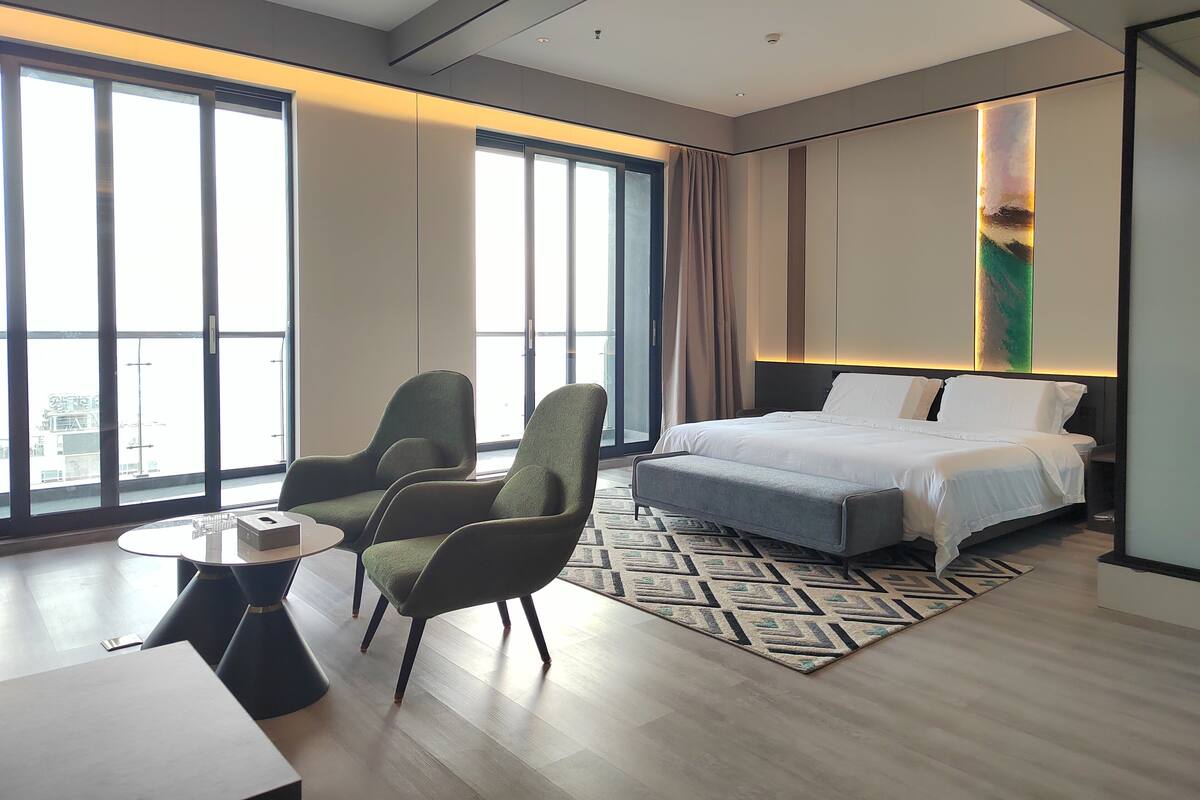 A modern hotel room with a large bed, two green armchairs, a small round table, and a view of the city through large windows.