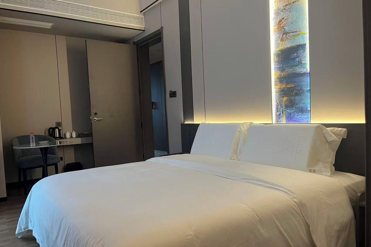 A modern hotel room with a large bed, a small desk with a chair, and a wall-mounted air conditioner.