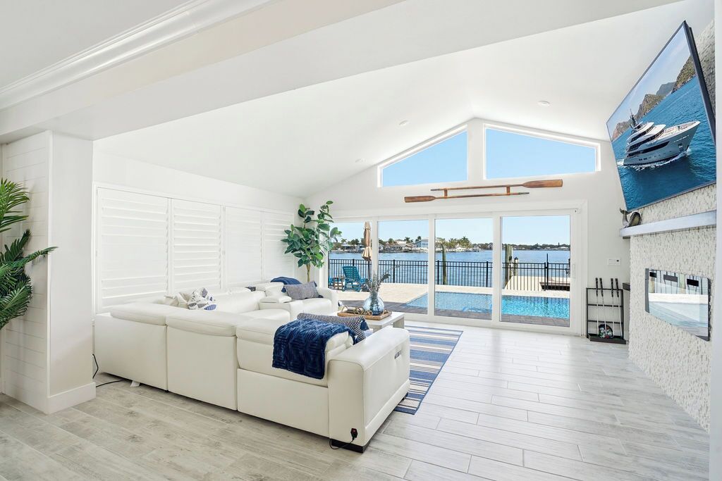 A spacious living room with a white sofa, a large window overlooking a pool and a yacht, and a high ceiling with skylights.