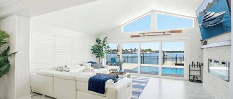 A spacious living room with a white sofa, a large window overlooking a pool and a yacht, and a high ceiling with skylights.