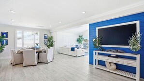 A modern living room with a dining area, a TV on a blue wall, and a white sofa. - Yacht Club Waterfront Home 4BR + Den & Pool (Indian Rocks Beach)