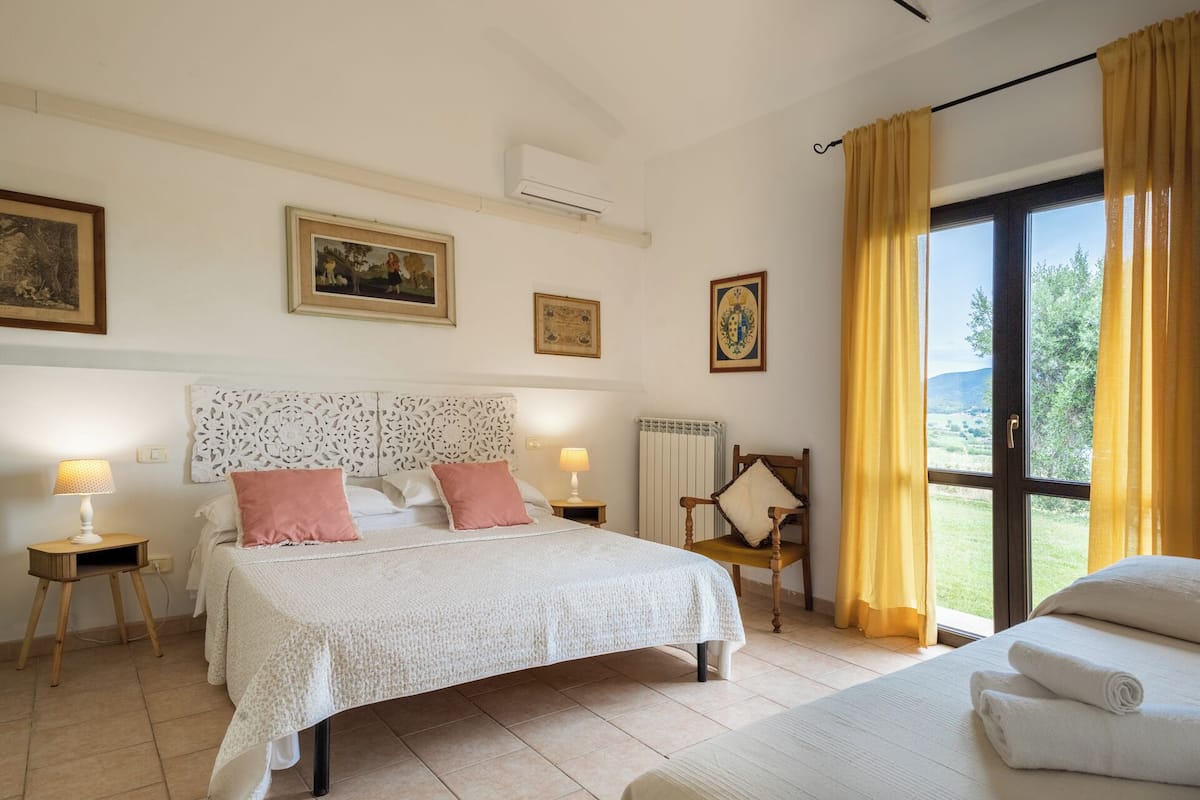 Une chambre à coucher comprenant un lit, des tables de chevet, une chaise, une fenêtre avec des rideaux et des œuvres d’art accrochées au mur.