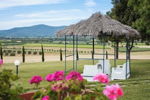 A gazebo with a thatched roof, white wicker furniture, and a view of a vineyard and hills. - Agriturismo il Montoto - Nature & Relax (Castiglione della Pescaia)
