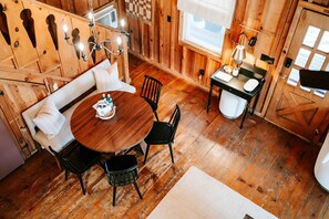 A cozy wooden cabin interior with a round wooden table, two black chairs, a bench with pillows, a small table with a lamp, and a window with a checkered curtain.