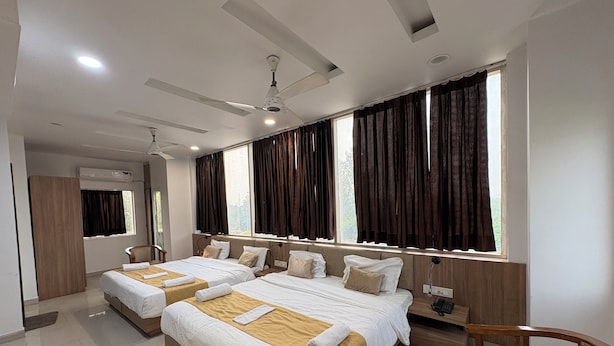 A hotel room with two beds, a ceiling fan, a large window with curtains, and a wooden chair. - Hotel Silver Bell (Lucknow)
