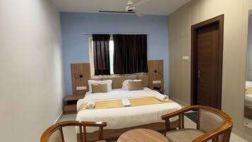 Habitación de hotel con una cama, dos sillas de madera, una mesita redonda, un ventilador de techo y una puerta.