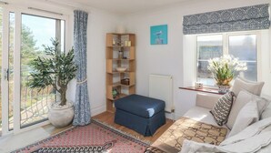 A cozy room with a large window, a blue sofa, a wooden bookshelf, and a potted plant. - 4 The Elms (Salcombe)