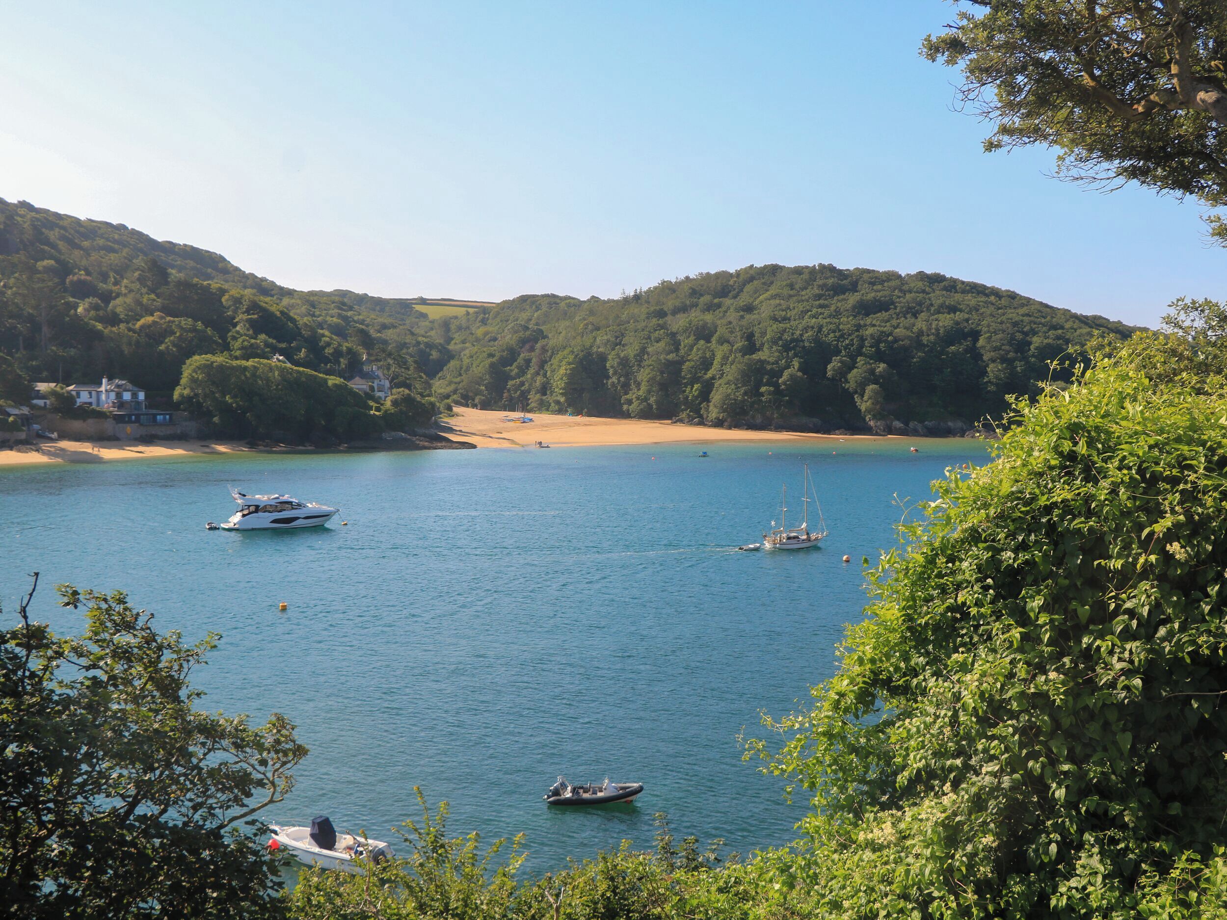 Un paysage côtier avec des bateaux sur une eau calme, entouré de collines verdoyantes et d’une plage de sable.