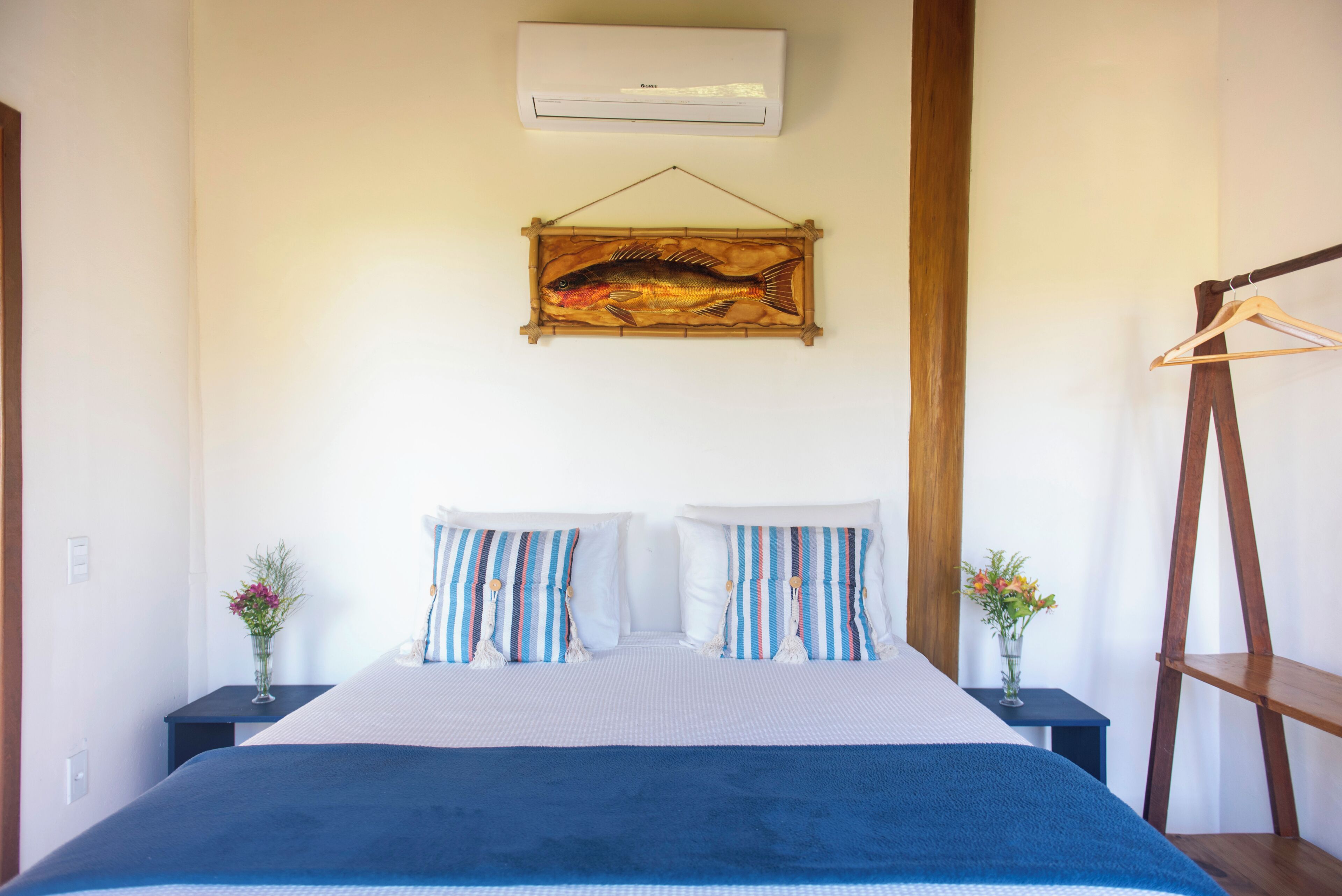 A bedroom with a bed, two bedside tables, a hanging fish painting, an air conditioner, and a wooden hanger.